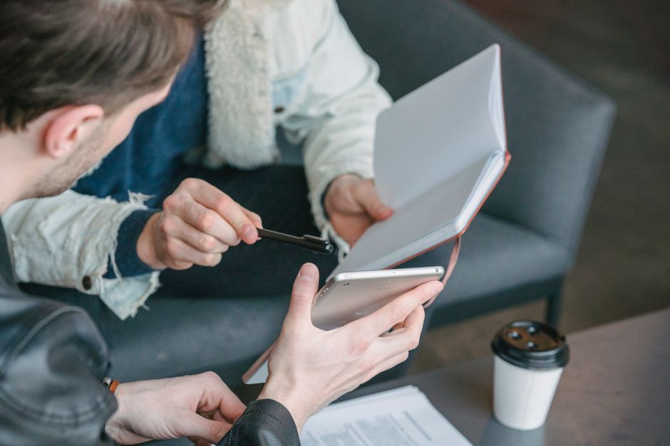 High angle of crop businessman showing tablet to male partner sitting on sofa with notepad while working together